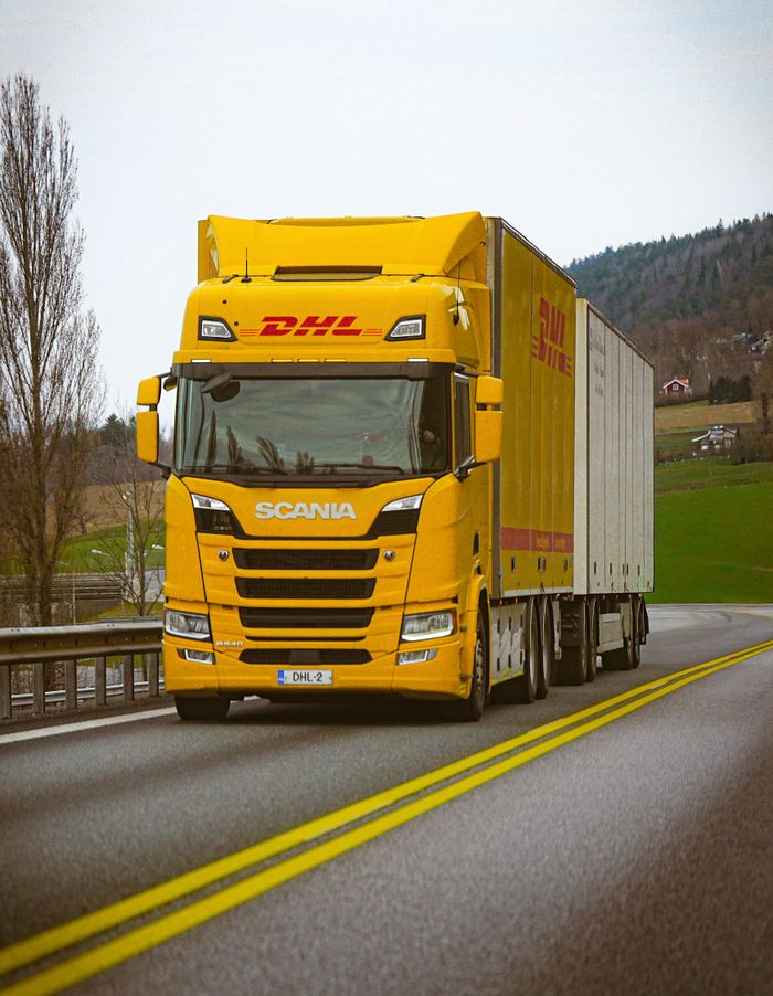 A yellow DHL truck on a scenic highway with green hills in the background.
